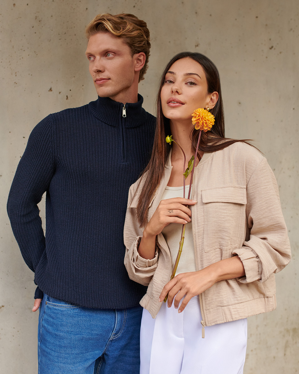 Homme en pull bleu marine et jean, femme en veste beige et pantalon blanc, tenant une fleur.