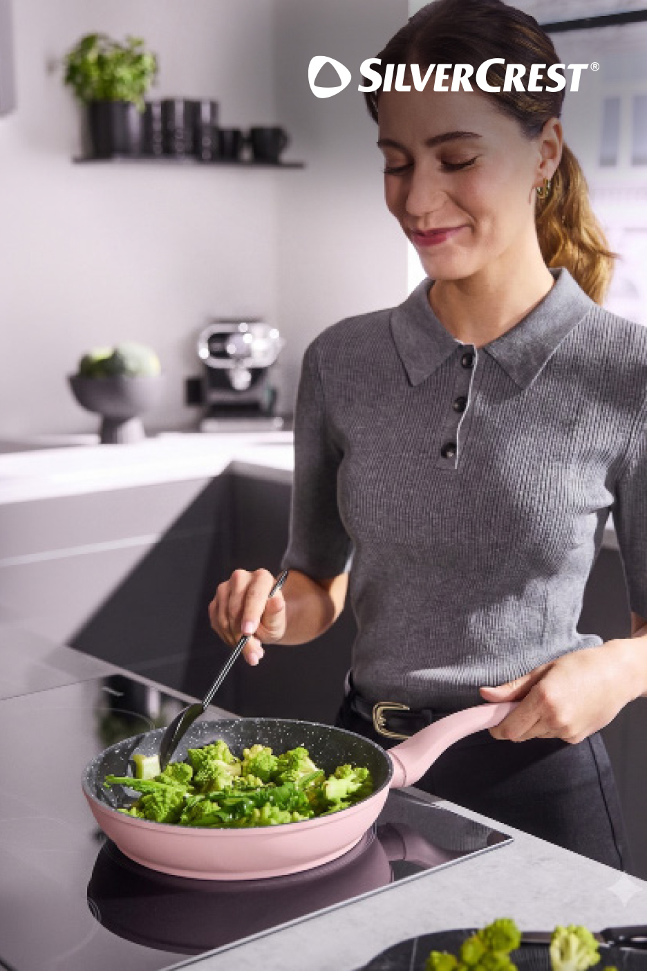 Une femme souriante cuisine des brocolis dans une poêle rose sur une plaque de cuisson à induction.