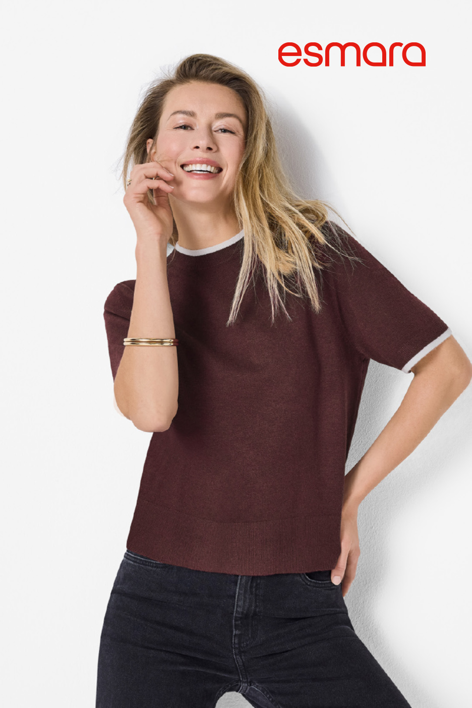Femme souriante portant un t-shirt bordeaux à manches courtes et un jean foncé.