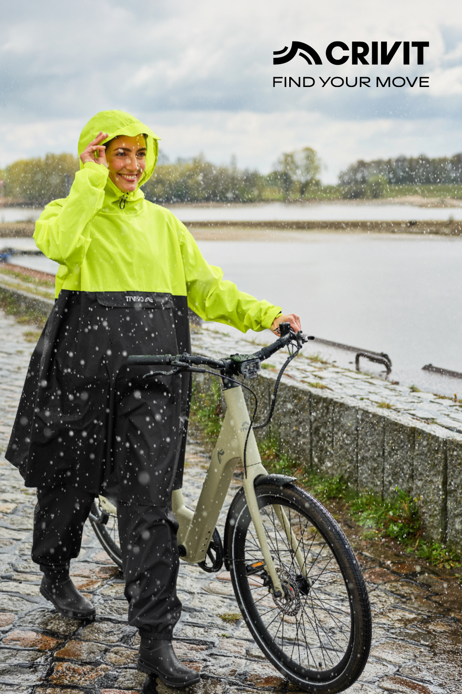 Femme souriante en poncho de pluie jaune fluo et pantalon noir, marchant avec un vélo sous la pluie.
