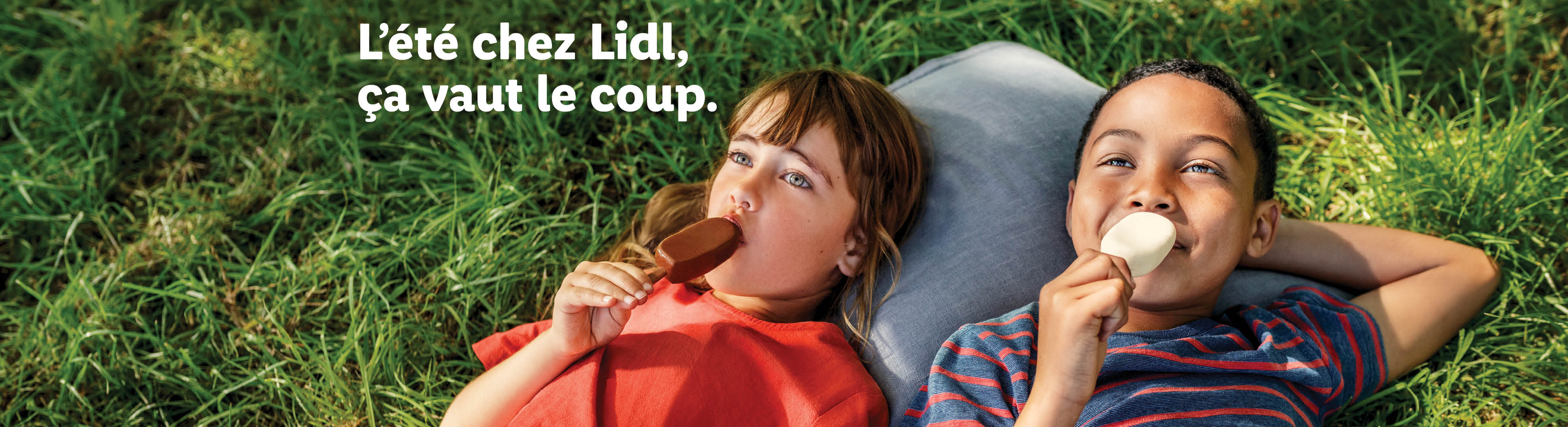 Deux enfants souriants mangent des glaces sur l'herbe. Texte: L'été chez Lidl, ça vaut le coup.