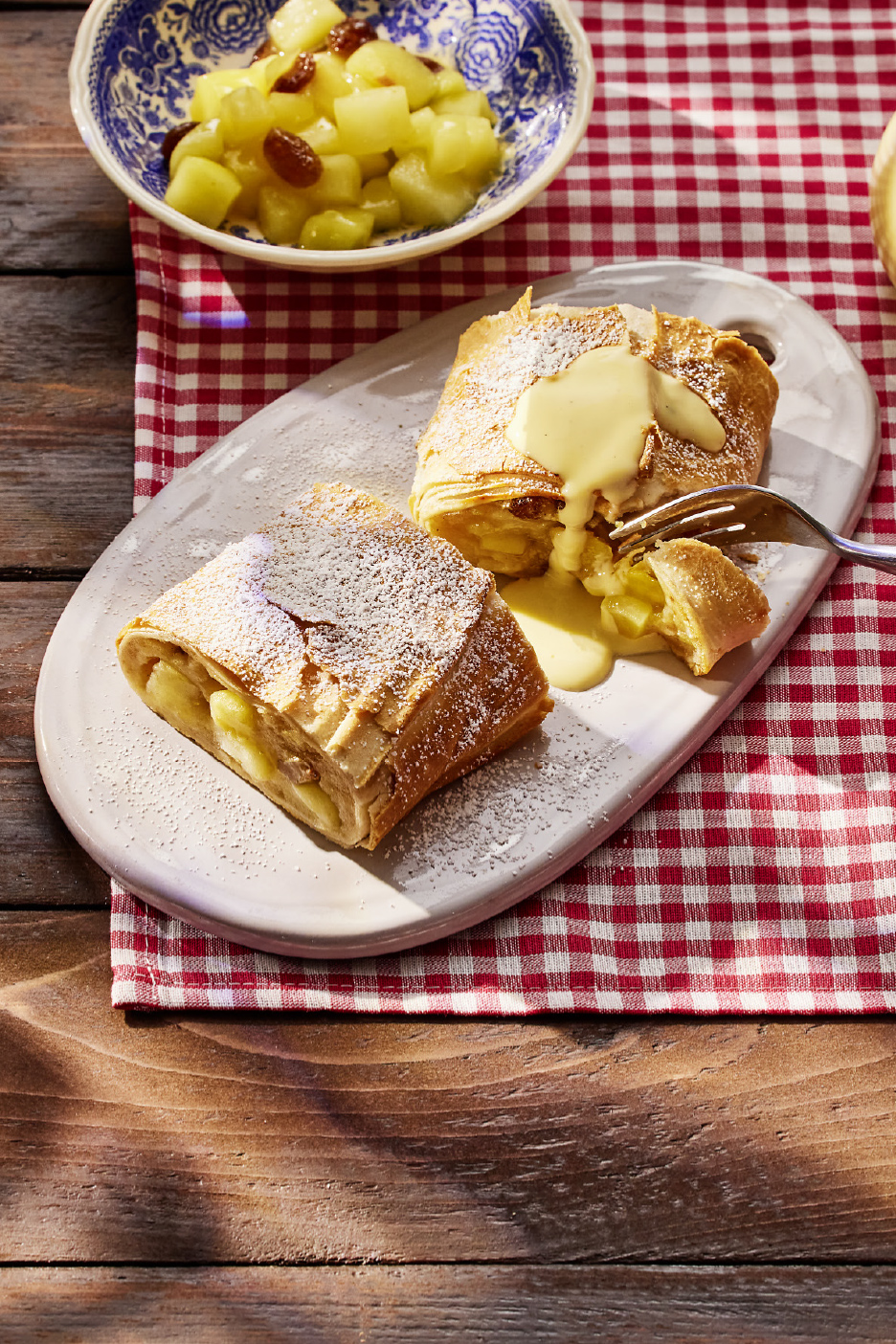 Strudel aux pommes avec sauce vanille et compote de pommes sur une nappe à carreaux.