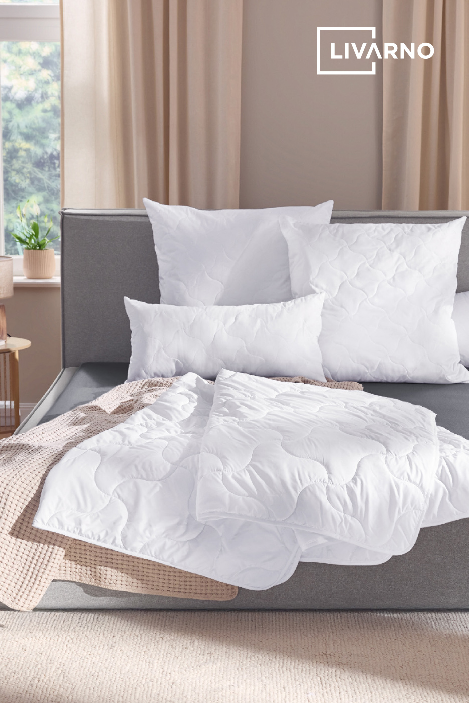 Literie blanche matelassée sur un lit gris dans une chambre lumineuse.