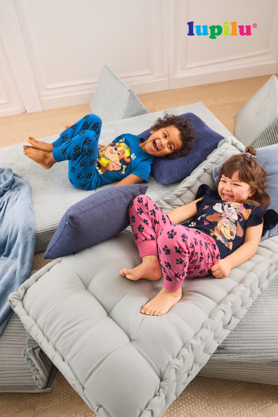 Deux enfants rieurs en pyjama à motifs de pattes de chien, allongés sur des coussins de sol.
