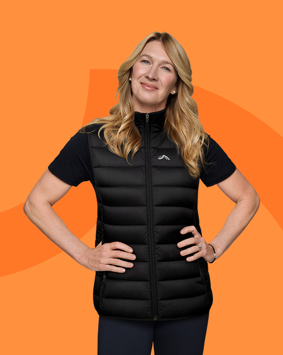 Femme en gilet matelassé noir et t-shirt, mains sur les hanches, souriant sur fond orange.