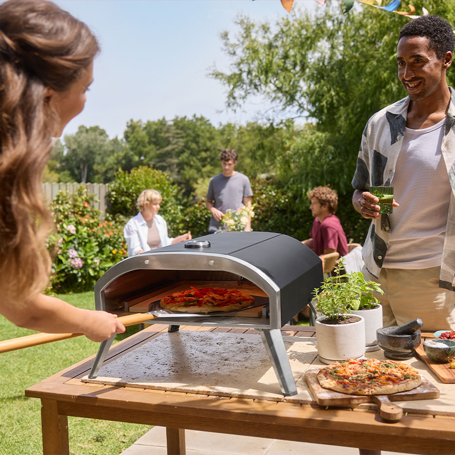 Famille à une fête de jardin, femme mettant une pizza dans un four, homme tenant une boisson.